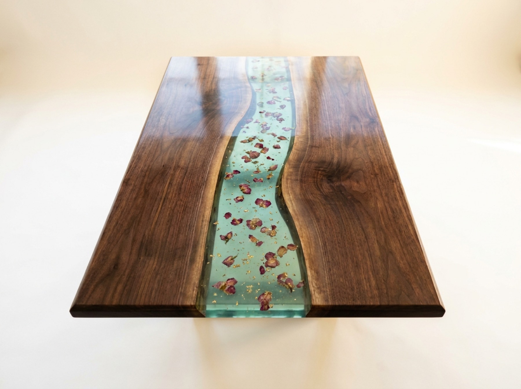 A walnut table top from above, with a wide sea-green resin river holding dried rose petals and gold flake.